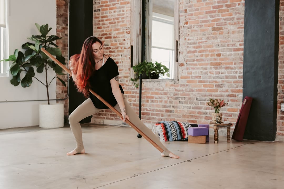 Krysta demonstrating a lunge with a wooden staff in a brick-walled movement studio